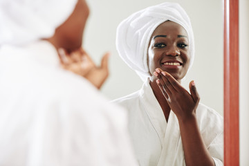 Horizontal shot of beautiful young Black woman standing in front of mirror applying nourishing cream on her face in morning