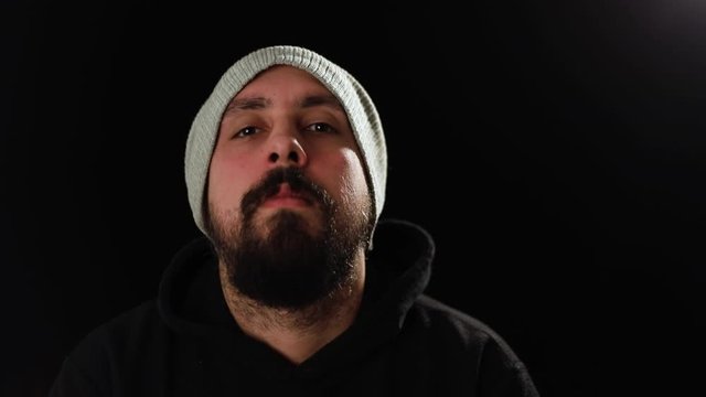 Thoughtful young man with beard and hat massaging his face and looking up, thinking in front of camera