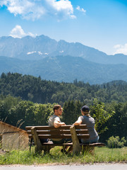 Fototapeta na wymiar A couple sits on a bench and enjoys a beautiful view of the Alps