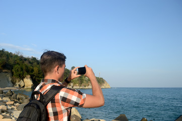 A young man, a tourist with a backpack, explores new places and shoot photos and makes a video of the summer seascape to share on social networks
