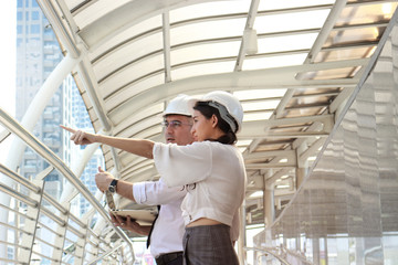 Fototapeta premium Two engineer wearing white safety helmet using a laptop together, male and female discussing about project work, pointing something, while walking on the way outside office in downtown city