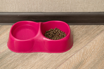 Double, pink plastic bowl for feeding cat stands on light laminate. Cells are filled with dry food and water for drinking. Concept of love and care for Pets
