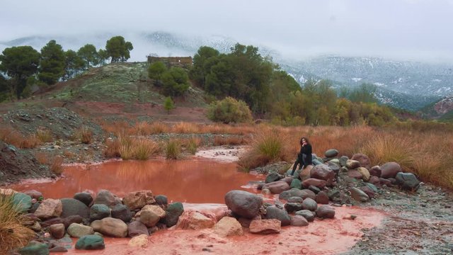 Cinemagraph / seamless video loop of a young female woman model sitting at a river with red water in the Atlas mountains in Morocco with snow on the peaks of the distant mountains.
