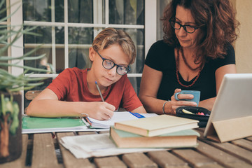 Young student doing homework at home with school books newspaper, digital pad helped by his mother. Mum control, help and teaching his son. Education, family, lifestyle and homeschooling concept.