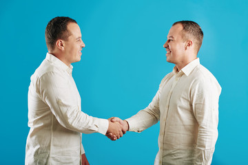 Horizontal side view medium studio portrait of twin brothers wearing white shirts shaking their hands, blue background