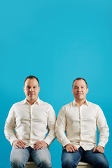 Vertical studio portrait shot of two twin brothers sitting on chairs against blue wall background looking at camera