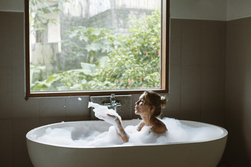Woman relaxing in foam bath with bubbles in dark bathroom by window
