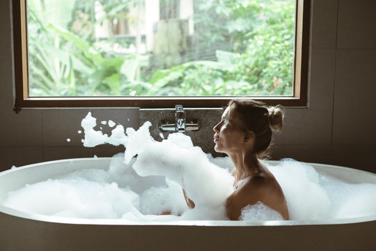 Woman relaxing in foam bath with bubbles in dark bathroom by window