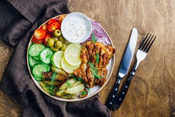Lunch bowl with grilled chicken, red onion, tomato, cucumber, lemon, olive and sause on wooden background. Top view.
