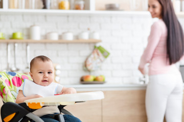 Baby playing on high chair in kitchen, mother washing dishes