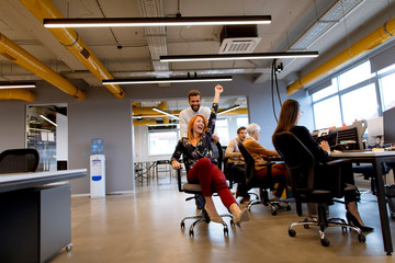 Young business people in smart casual wear having fun while racing on office chairs and smiling