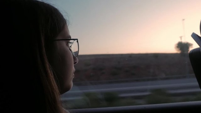 Pensive young woman thinking and looking out from train or bus window.  Casual brunette with glasses in sharp, dim natural light.