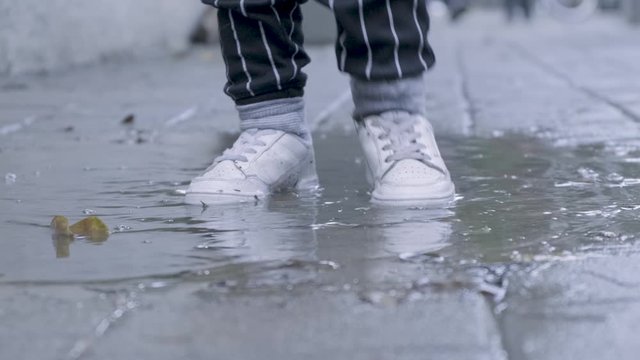 Cute baby girl with tiny white sneakers jumping and playing in a muddy puddle of rain water. cold wet winter 4k