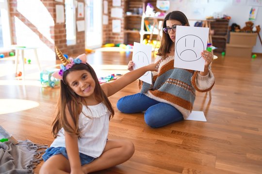 Beautiful pshychologist and toddler doing therapy with emotions emoji around lots of toys at kindergarten