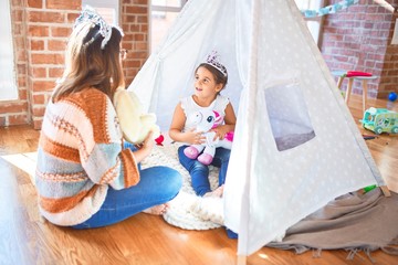 Beautiful teacher and toddler wearing princess crown playing with unicorn doll inside tipi around...