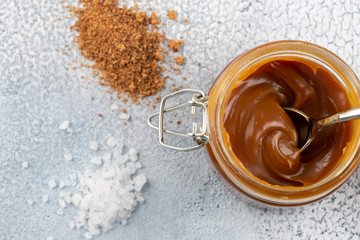 Glass jar with salted caramel and spoon on light background, copy space