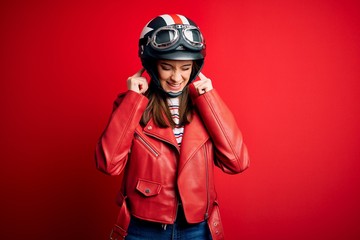 Young beautiful brunette motocyclist woman wearing motorcycle helmet and red jacket covering ears...