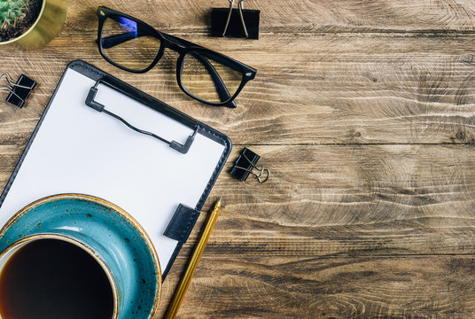 Office table desk. Workspace with blank clip board, office supplies, pen, glasses, green leaf and coffee cup on wooden background. Flat lay, top view