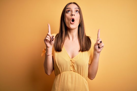 Young beautiful brunette woman wearing casual dress standing over yellow background amazed and surprised looking up and pointing with fingers and raised arms.