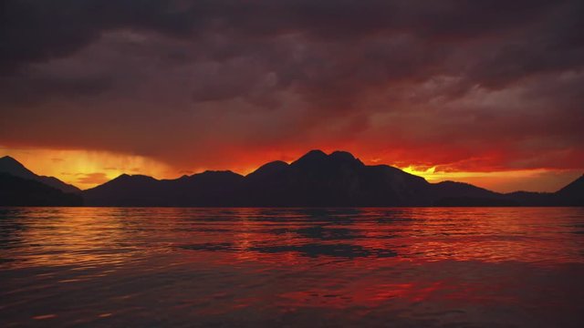 Scenic time-lapse cinemagraph / seamless video loop of a beautiful and romantic sunset at mountain lake Walchensee in Southern Germany / Bavaria with moving red clouds and calm waves in real time.