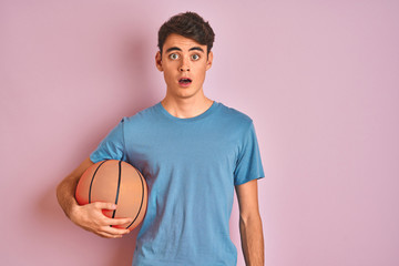 Teenager boy holding professional basket ball over isolated pink background scared in shock with a surprise face, afraid and excited with fear expression