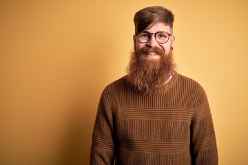 Handsome Irish redhead man with beard wearing glasses and winter sweater over yellow background...