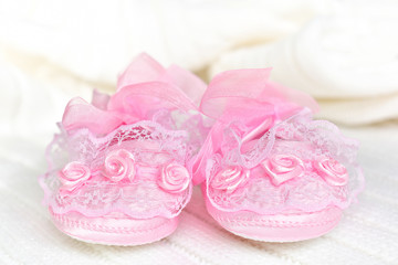 Fototapeta na wymiar Pink baby booties from the front, on white crochet blanket. Shoes with lace, shiny ribbon and silk rose decoration.