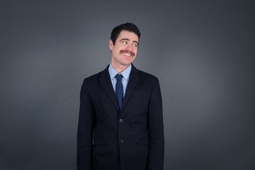 Dreamy young male with thoughtful expression, looks away keeps hands down bitting his lip thinks about something pleasant poses against gray wall.