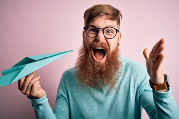 Irish redhead man with beard holding paper plane over pink isolated background very happy and excited, winner expression celebrating victory screaming with big smile and raised hands