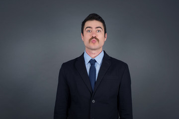 Attractive light haired male rounds lips, wears casual clothes, looks directly into camera, expresses her satisfaction, isolated over gray background. People, body language conceptâ€ 