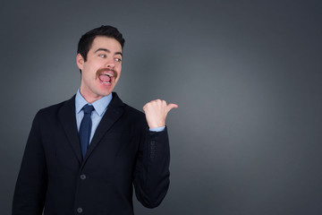 Omg concept. Stupefied caucasian male with surprised expression, opens eyes and mouth widely, points aside with thumb, shows something strange on gray background. Advertisement concept.