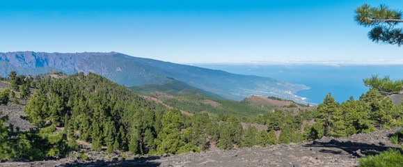 Panoramatic view of volcanic landscape with lush green pine trees, colorful volcanoes and white clouds at path Ruta de los Volcanes, hiking trail. La Palma, Canary Islands, Spain, Blue sky background © Kristyna