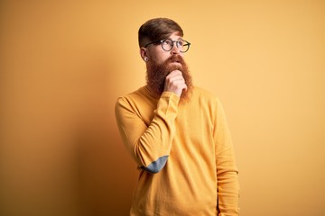 Handsome Irish redhead man with beard wearing glasses over yellow isolated background with hand on chin thinking about question, pensive expression. Smiling with thoughtful face. Doubt concept.