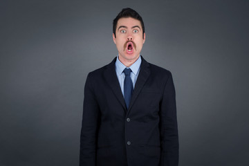 Emotional businessman with opened mouth expresses great surprisment and frighteness, poses against white concrete background, stares at camera. Unexpected shocking news and human reaction.