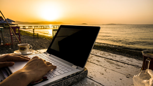 Freelance female hands working with laptop on wooden table on beach at sunset. Woman sitting on chair and using computer near blue sea shore at sunny day. Digital nomadism towards yellow sky & horizon