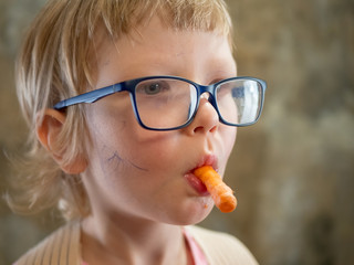 funny little blond boy in pink sweatshirt, big glasses and corset to correct posture eating fresh carrot. concept of vegetarianism and proper balanced nutrition for children