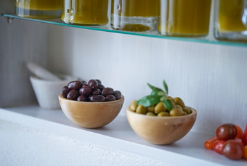 Atmospheric close-up picture of green and black olives in wooden bowls next to blurred white mortar and tomatoes, glass bottles with oil content can be seen in the cut  wall shelf stands in a Mediterr