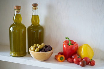 two wonderful bottles of cooking oil or olive oil on a wall shelf, wooden bowl with olives green and black next to vegan vegetarian vegetables such as tomatoes and peppers in spain