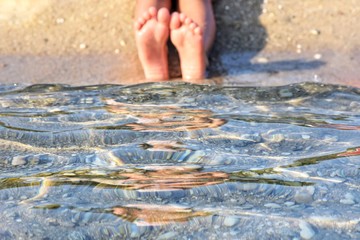 Soft focus. Girls bare feet at the transparent sea water with golden sand, selective focus. Bare feet in a sea wave.  Relaxation in the ocean.  Summertime. Barefoot girl plays with waves 