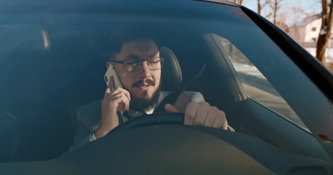 Young Caucasian handsome businessman in glasses speaking on the mobile pone while driving his car on the way to work in the morning,