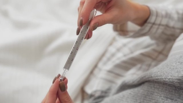 Close-up scene of a girl who is lying in the bed and holding a mercury thermometer in her hand. She has a fever.
