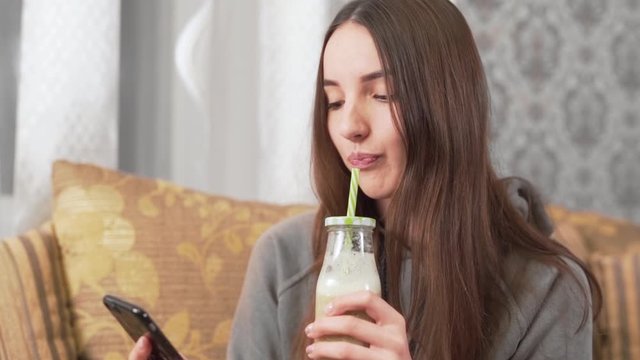 Beautiful girl is sitting at home on the couch, drinking a homemade milkshake from a bottle with a straw and clicking in a phone.