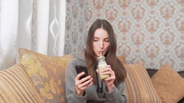 Beautiful girl is sitting at home on the couch, drinking a homemade milkshake from a bottle with a straw and clicking in a phone.