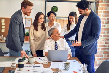 Group of business workers smiling happy and confident. One of them sitting and partners standing around. Working together with smile on face looking at the laptop at the office