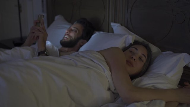 beautiful young couple laying in bed, guy playing with his phone and laughing while girl is annoyed trying to sleep. 4K