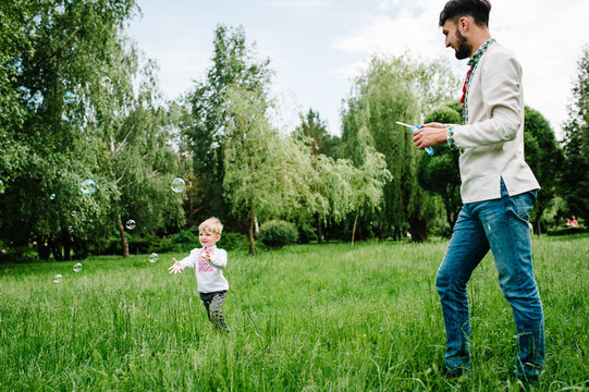 Happy Little Baby Boy Running In Embroidered Shirt On The Background Of Nature Catch Inflatable Bubbles. Dad Lets Out Bubbles For Her Son. Full Length. Looking Sideways. Close Up.