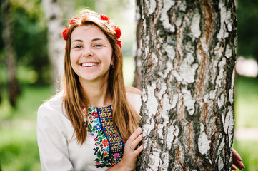 Young happy woman stands with a crown from flowers on the head, on the background of birches on the nature in an embroidered shirt, a embroidery handmade. Close up. upper half, looking at camera.