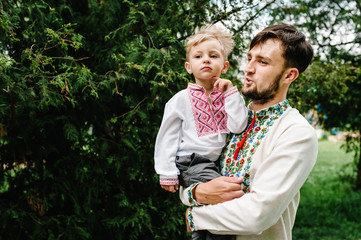 Angry little baby boy in funny dad on his hands in an  embroidered shirt, stand and poses on the background of nature. upper half. looking sideways. Close up.