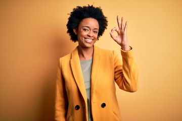 Young beautiful African American afro businesswoman with curly hair wearing yellow jacket smiling positive doing ok sign with hand and fingers. Successful expression.