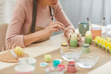 Fototapeta na wymiar Cropped portrait of unrecognizable young woman painting eggs in pastel colors for Easter while sitting at table in kitchen or art studio, copy space